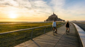 View of the Mont Saint-Michel tidal island in Normandy, France, at sunset with a couple biking to the town on the wooden jetty.