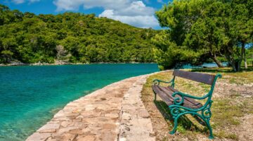 A rustic wooden and cast iron bench sits empty beside a stone walking path on St. Mary&#039;s Island in beautiful Veliko Jezero Lake, in Mljet National Park, Croatia.